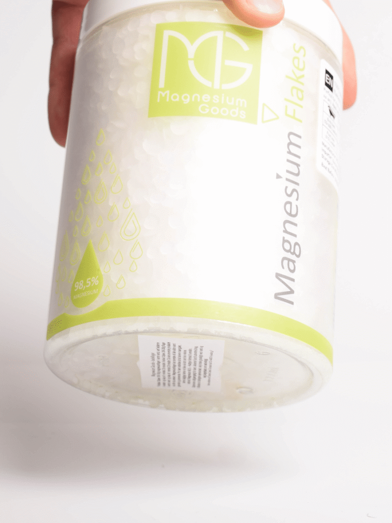 Hånd holder beholder med Magnesium Goods Magnesium Flakes, hvide flager og grøn etiket