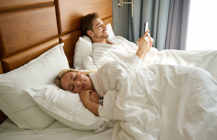 Mand i badekåbe ligger i stor seng og tjekker mobil, kvinde sover ved siden af under hvidt dyne