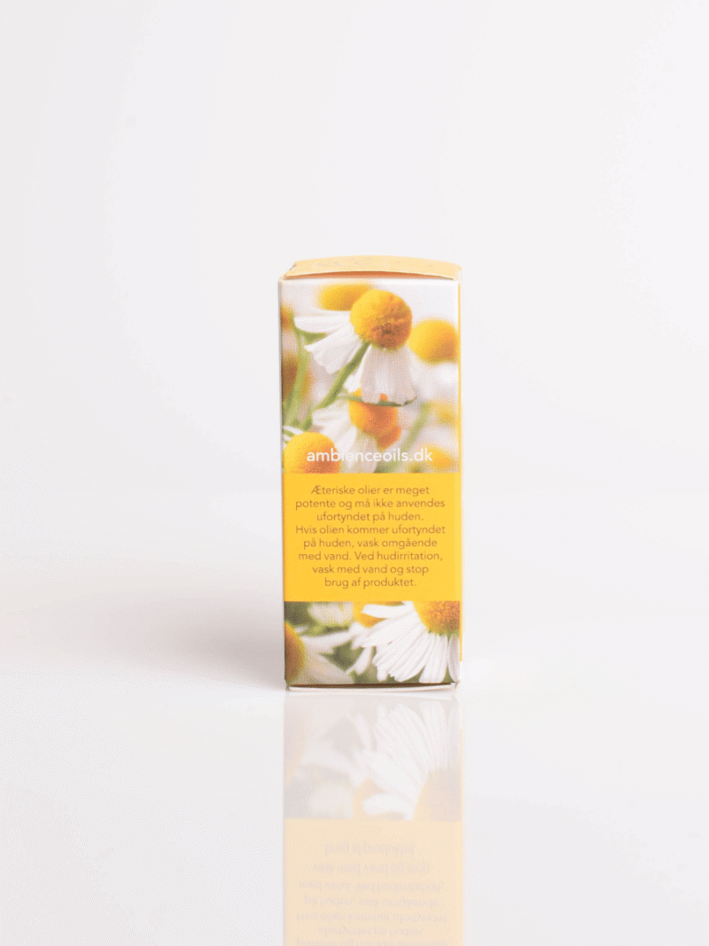 Gul æterisk olie-æske med kamilleblomster og dansk tekst stående på hvid, reflekterende overflade