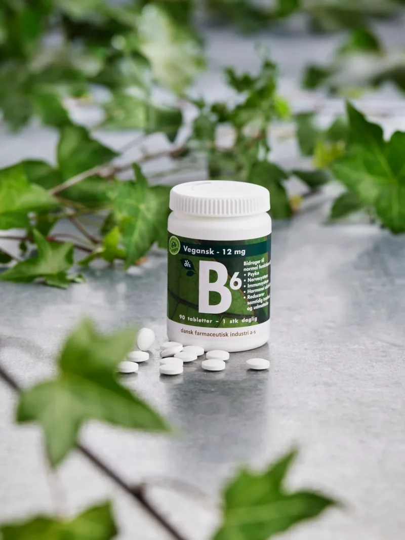 Flaske med B6 vitamintabletter, hvide piller spredt foran og grønne blade i baggrunden