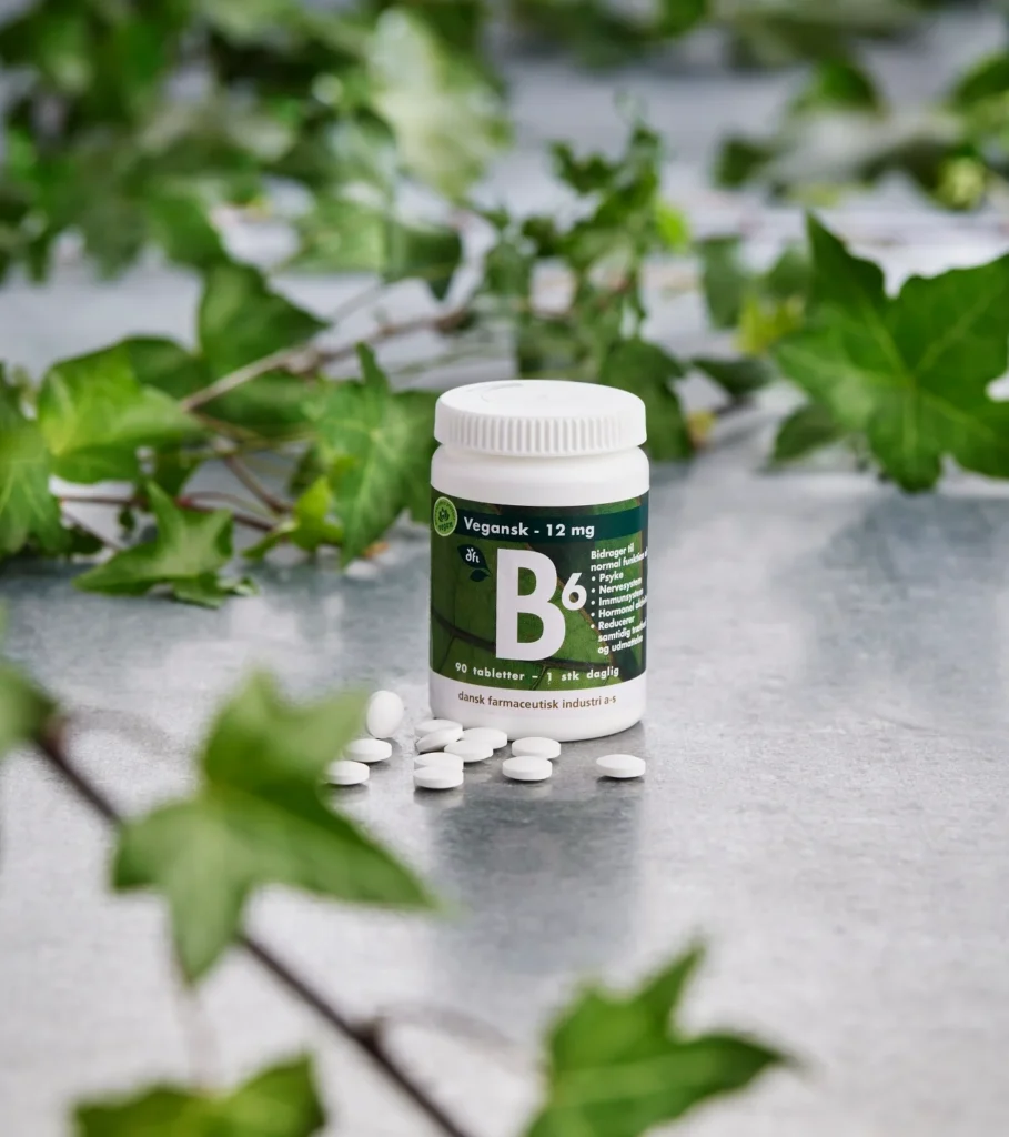 Flaske med B6 vitamintabletter, hvide piller spredt foran og grønne blade i baggrunden
