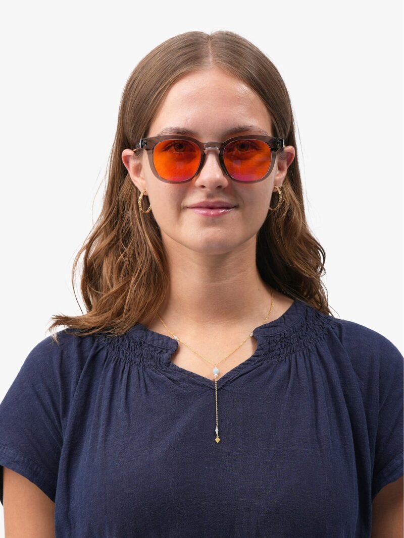 Kvinde med grå runde solbriller med orange spejlglas, blå bluse og gyldne smykker
