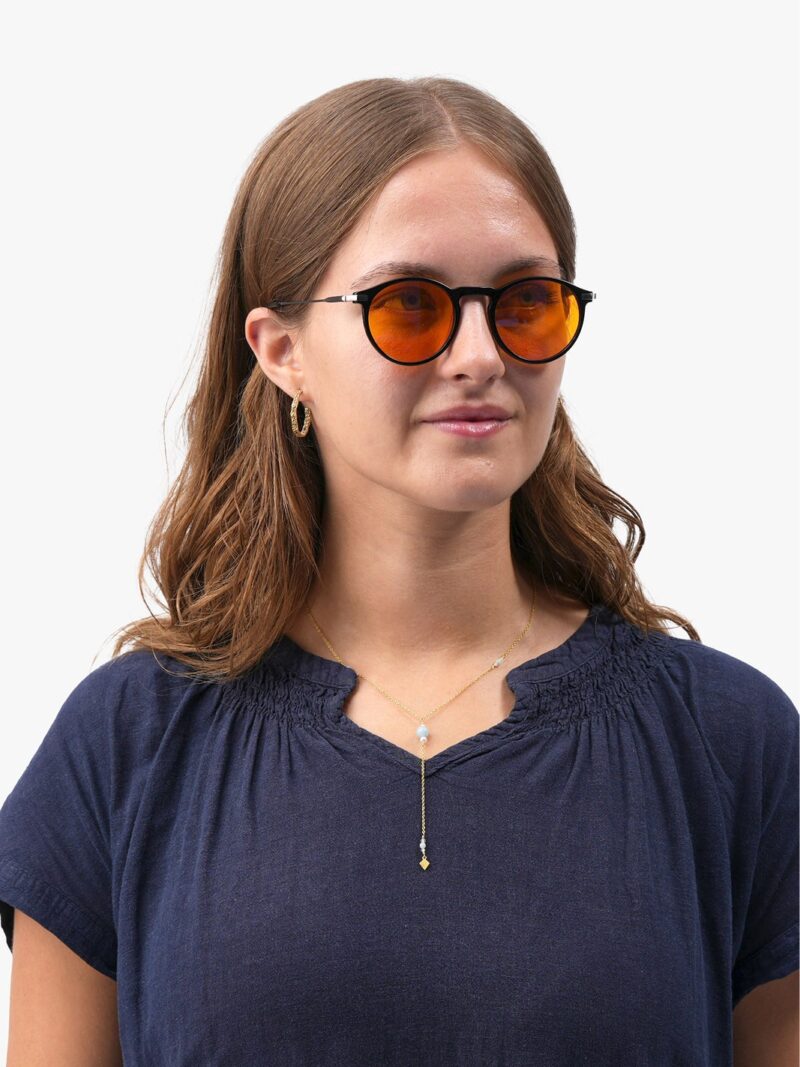 Kvinde med runde solbriller med orange glas, guld hoop-øreringe og delikat lariat-halskæde iført mørkeblå bluse