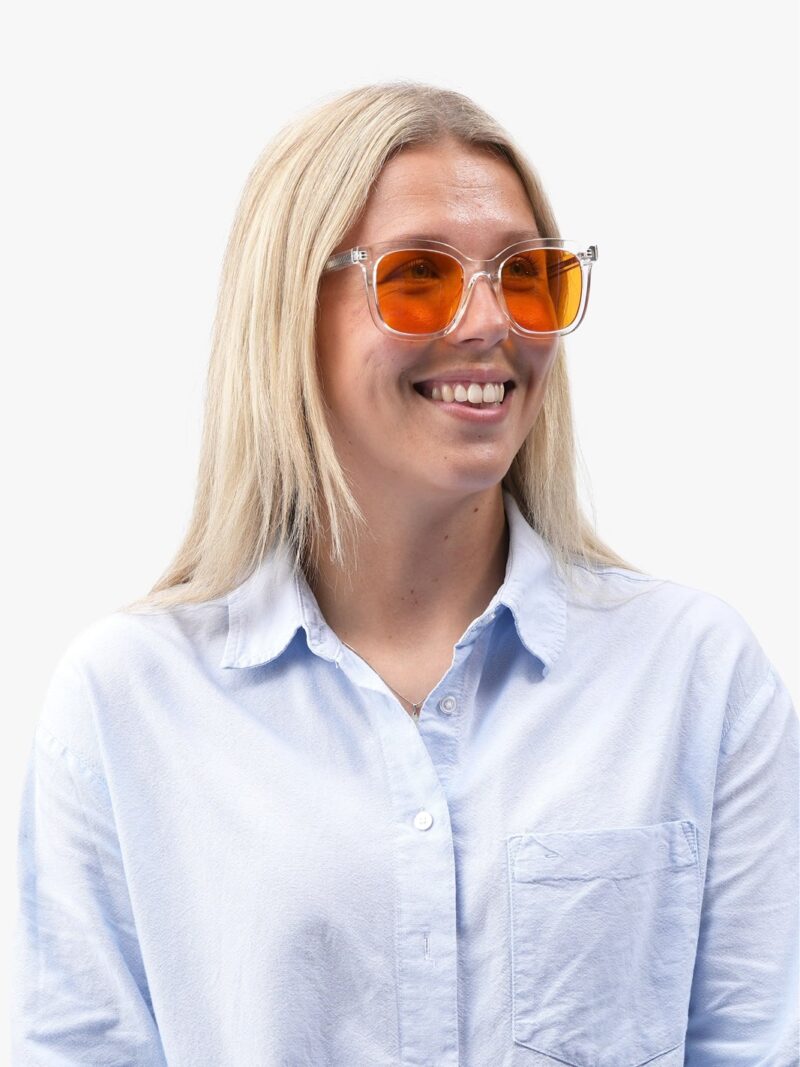 Smilende kvinde med blondt hår iført gennemsigtige solbriller med orange linser og lyseblå skjorte
