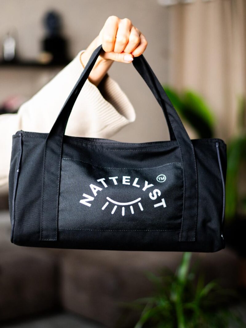 Sort stoftaske med NATTELYST-logo holdt i ét håndtag, nærbillede foran sløret stuebaggrund