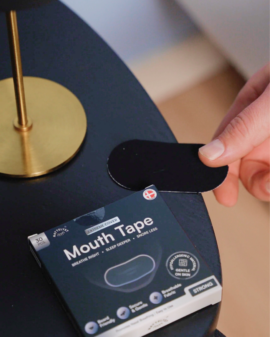 Hånd holder sort mundtape ved siden af æske med Mouth Tape på natbord