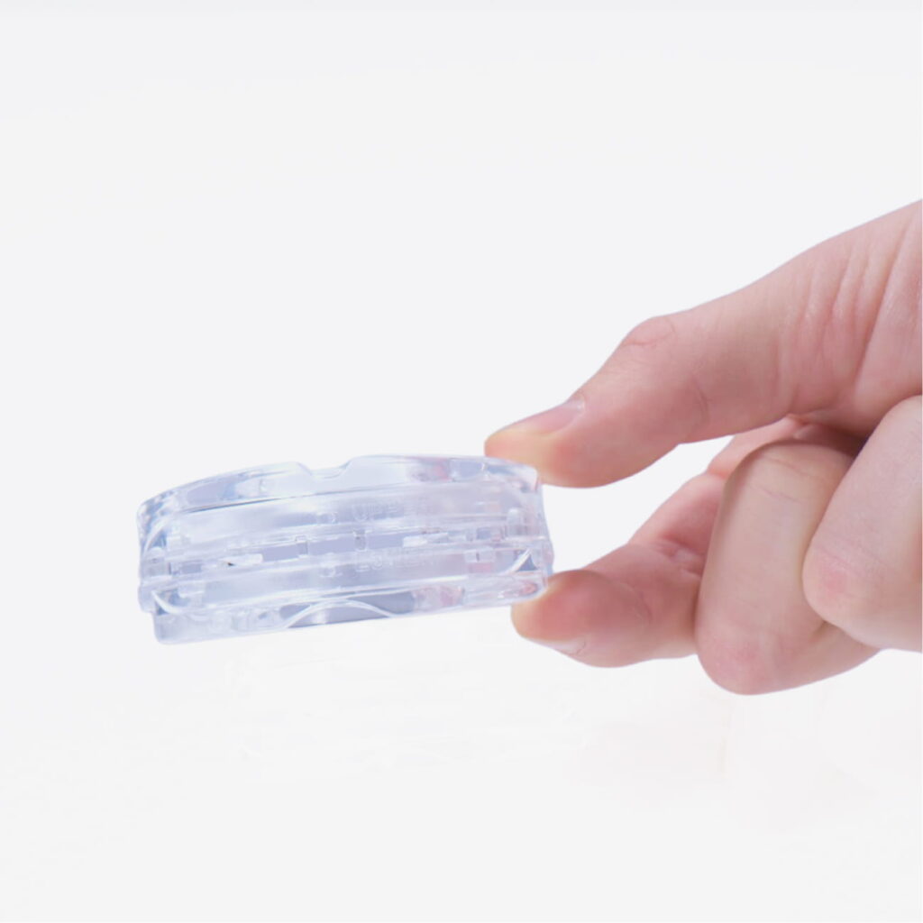 Hånd holder lille rund gennemsigtig plastkassette med indvendige rum og rille, på hvid baggrund